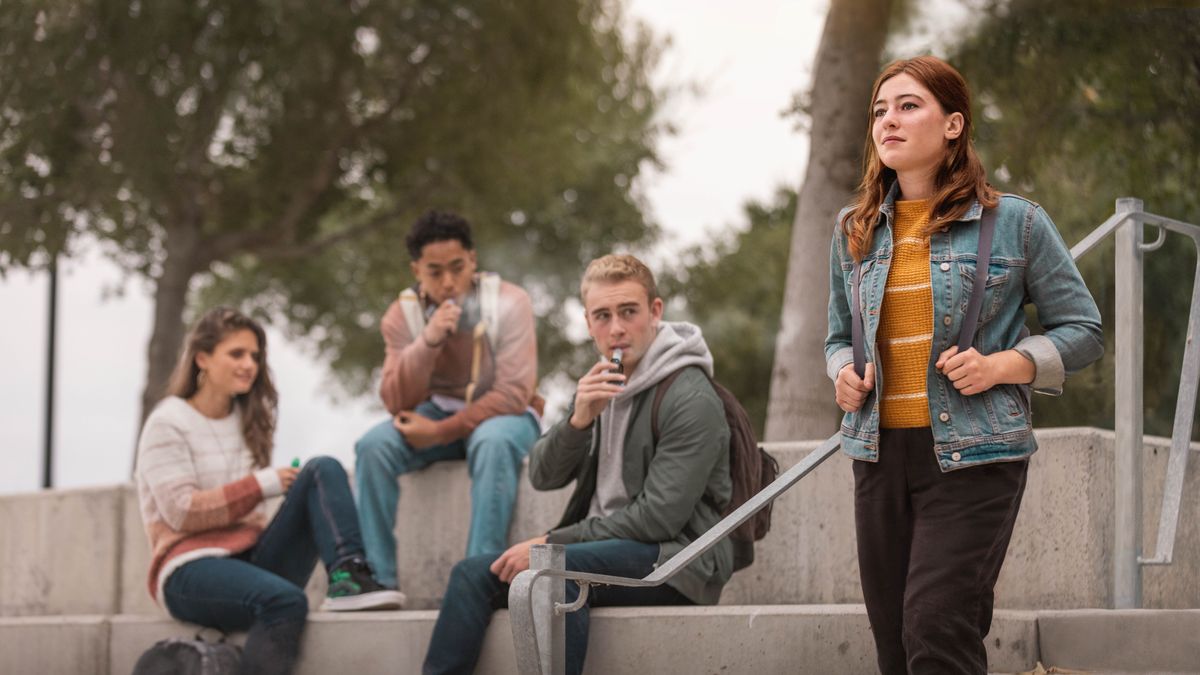A young sister walking home from school past her peers who are smoking electronic cigarettes. They stare at her as she confidently looks ahead.