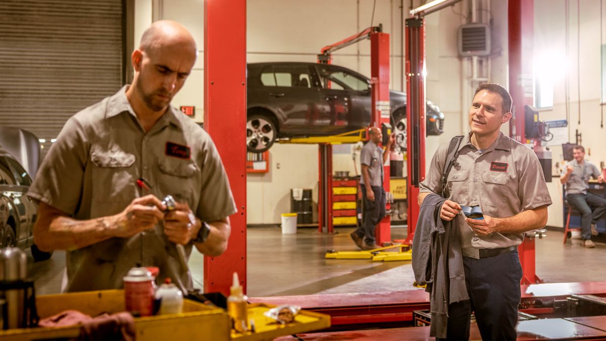 A brother working at an auto repair shop and approaching a tough-looking workmate to give him an invitation to a meeting. The brother’s other workmates are laughing at him.