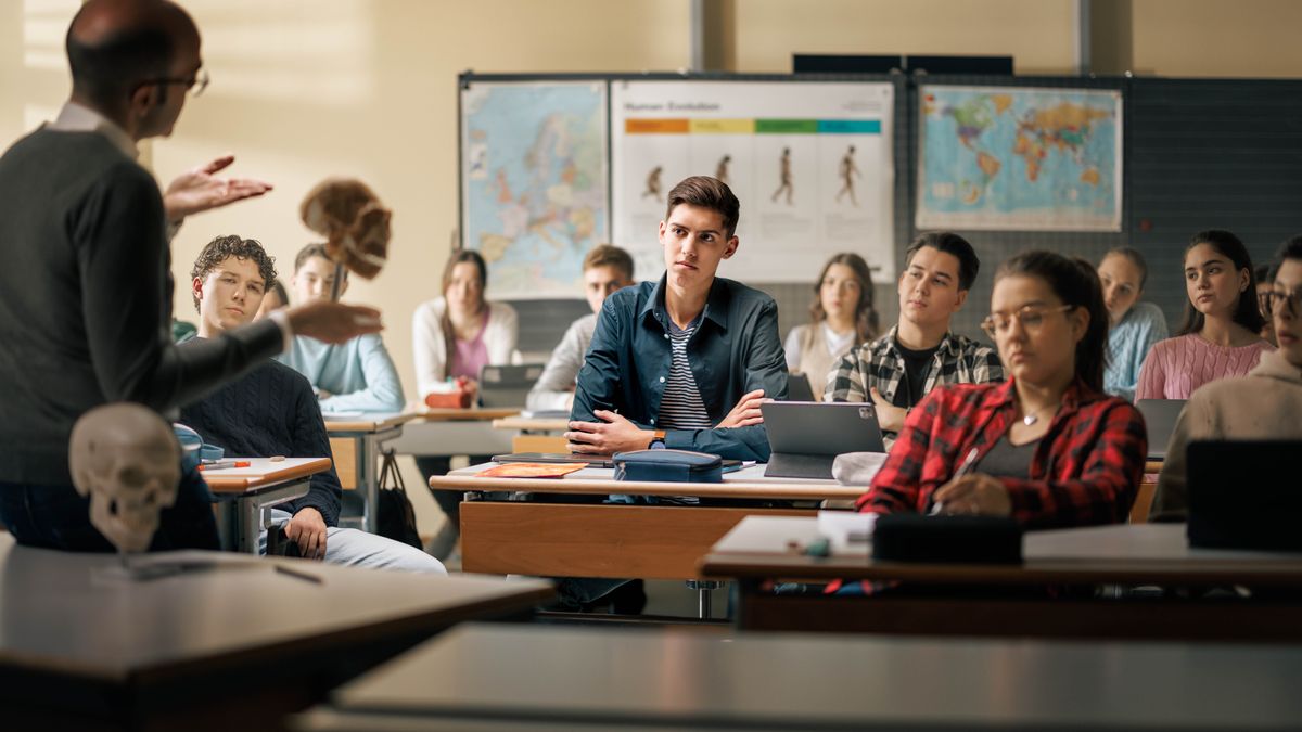 A teenage boy in a classroom listening respectfully as a science teacher gestures toward a skull and addresses the boy directly. At the back of the classroom is a conceptual image of the stages of human evolution.