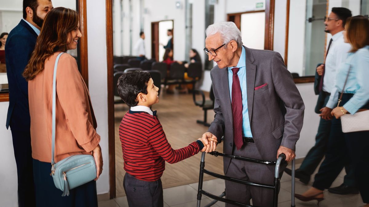 An elderly brother using a walker, joyfully shaking a young brother’s hand as he and his parents enter a Kingdom Hall.