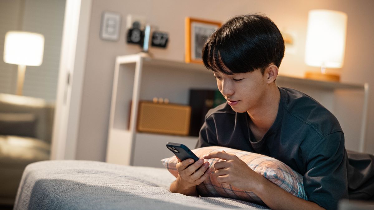 A young brother lying in bed staring at the screen of his cell phone.