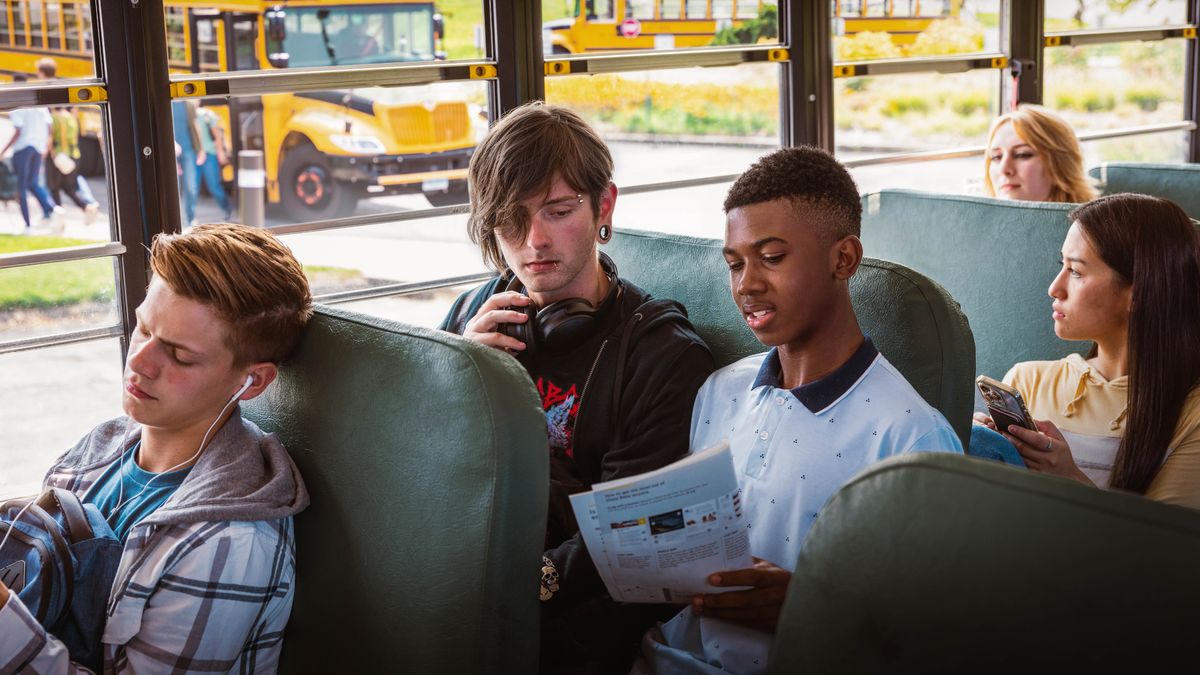 A teenage brother witnessing to a schoolmate on a bus. The schoolmate has long hair draped over part of his face and has piercings on his eyebrow, lip, and ear.