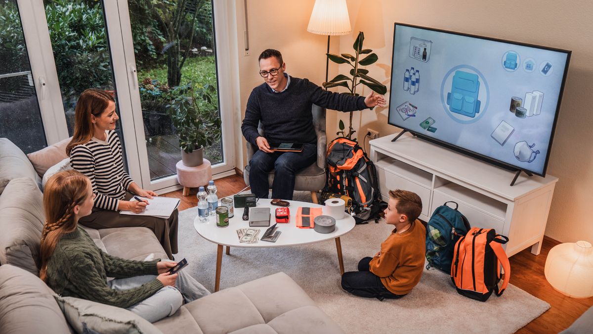 A father conducting family worship and helping his wife and two children prepare for potential disasters. They are watching the video “Are You Prepared for a Disaster?” Before them are bottles of water, cash, a Bible, canned food, duct tape, emergency go bags, and other items.