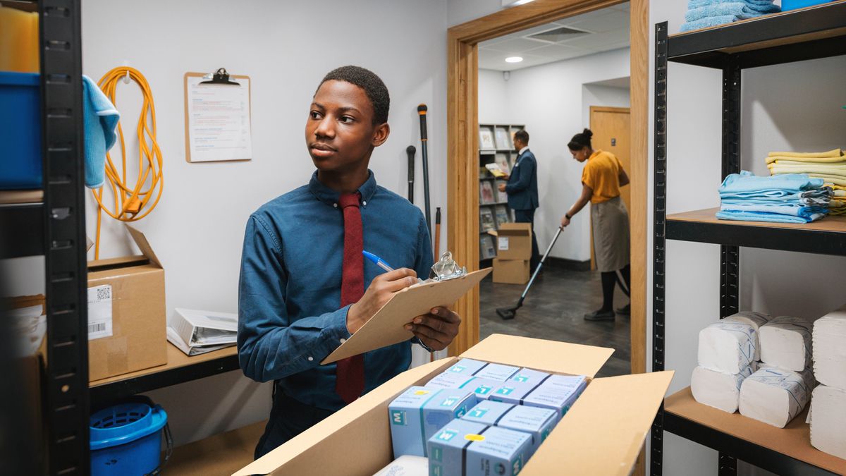 A teenage brother taking inventory of cleaning supplies at the Kingdom Hall. In the lobby, a brother restocks literature and a sister vacuums the floor.
